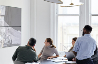 4 people around a table in a meeting or break room watching a wall-mounted TV screen.